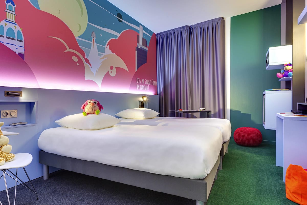 Une chambre d’hôtel avec un lit, un bureau, une chaise, un pouf rouge et une fresque murale.