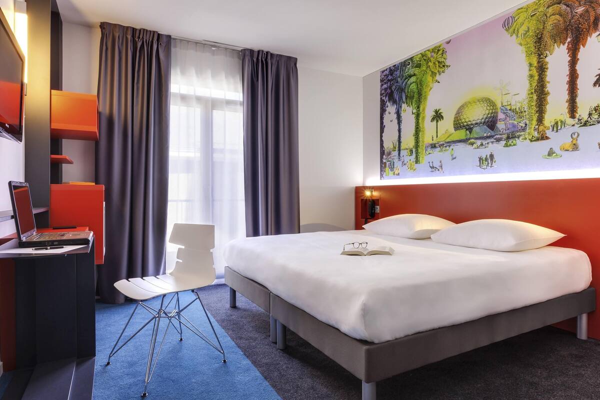 Una habitación de hotel moderna con una cama grande, un escritorio con una computadora portátil, una silla y una pared con un mural colorido.