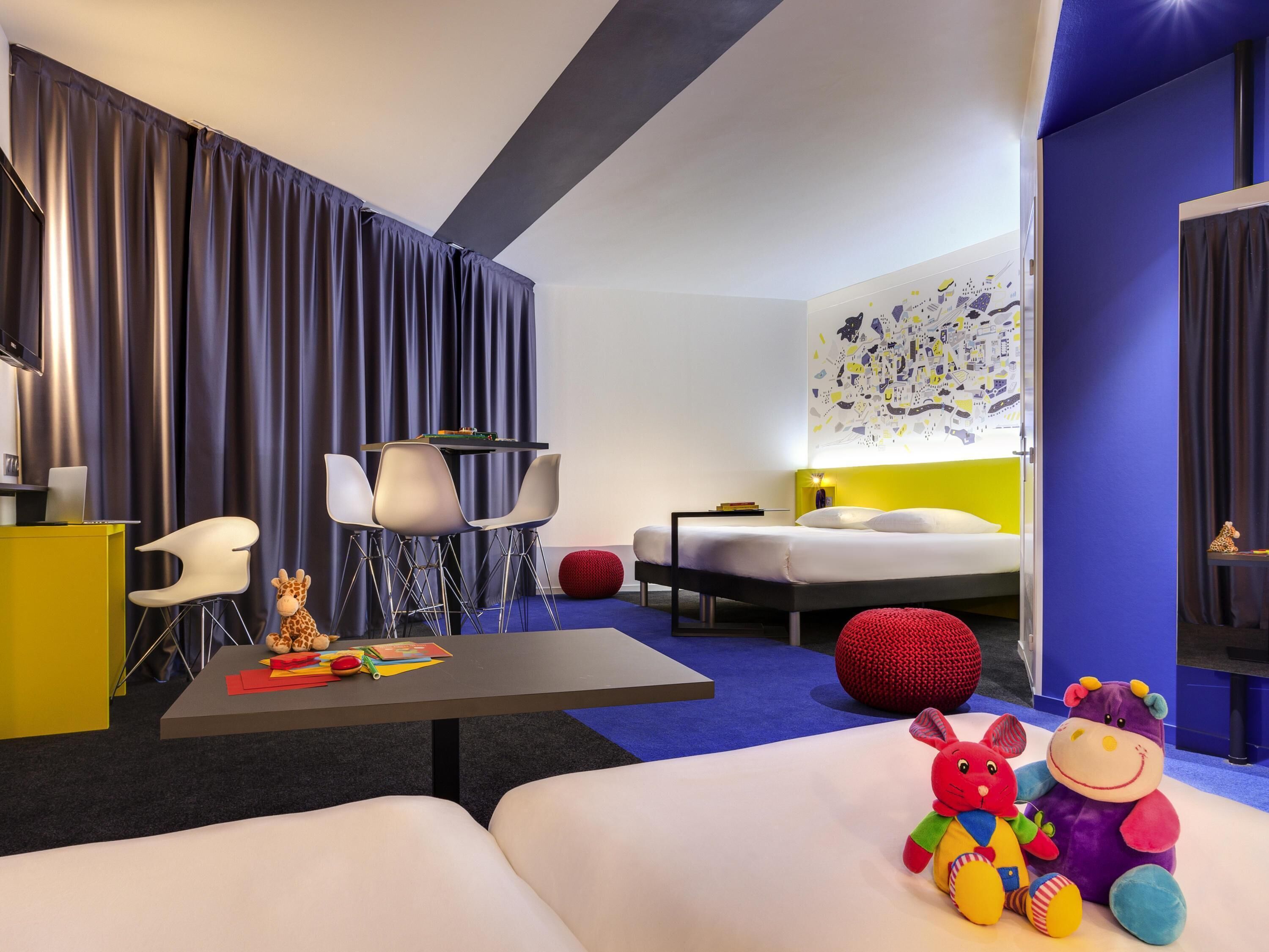 Una habitación de hotel moderna con una cama, un escritorio con una computadora portátil, una silla, una pelota roja y peluches.