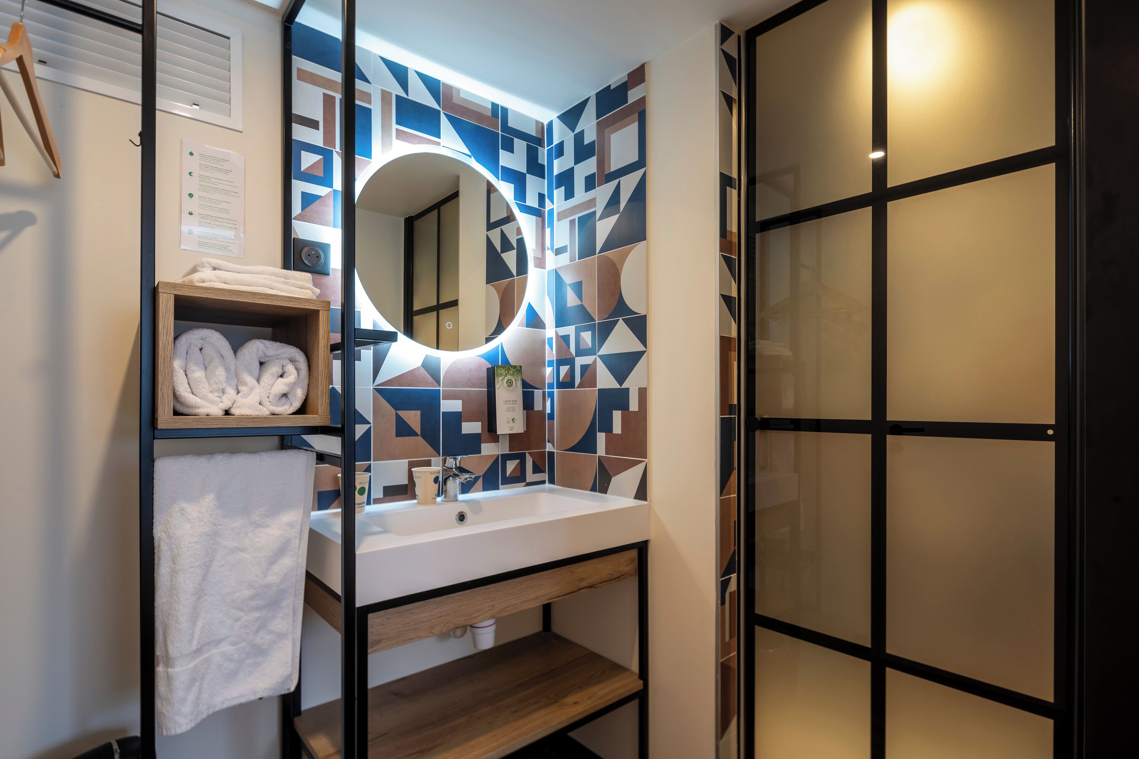 A modern bathroom with a geometric tile backsplash, a round mirror, a white sink, and a wooden shelf with towels.