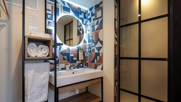 A modern bathroom with a geometric tile backsplash, a round mirror, a white sink, and a wooden shelf with towels.