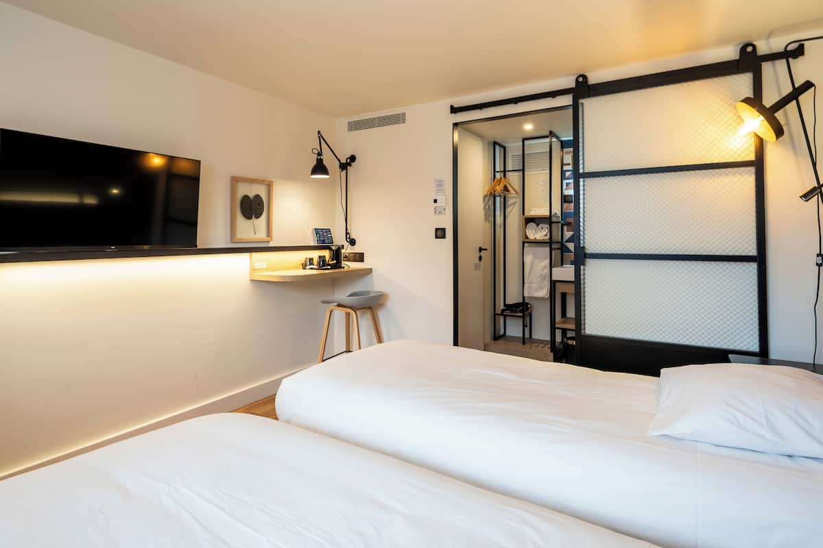 A hotel room with a large flat-screen TV, a desk with a lamp, a bed with white linens, and a sliding closet door.