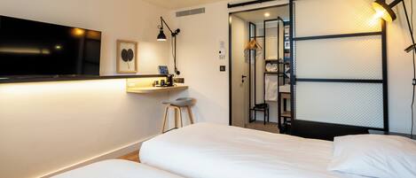 A hotel room with a large flat-screen TV, a desk with a lamp, a bed with white linens, and a sliding closet door.