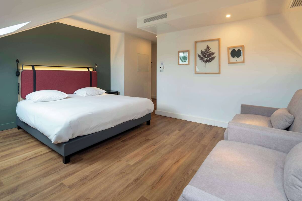 A bedroom with a bed, a sofa, wooden flooring, and wall-mounted artwork.