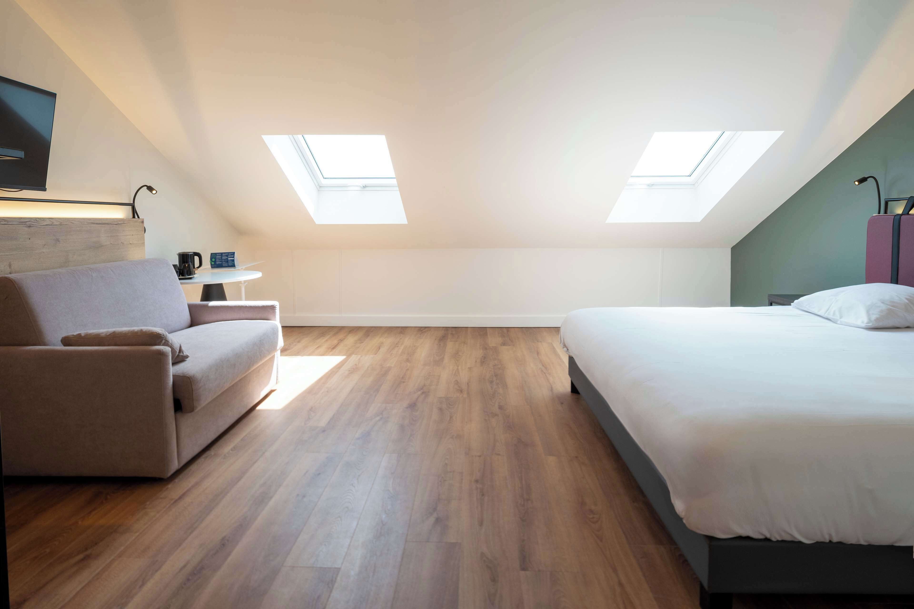 A modern, well-lit room with a bed, a sofa, and a small table. There are skylights on the ceiling, allowing natural light to enter.