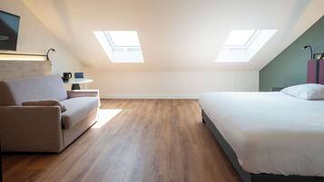 A modern, well-lit room with a bed, a sofa, and a small table. There are skylights on the ceiling, allowing natural light to enter.