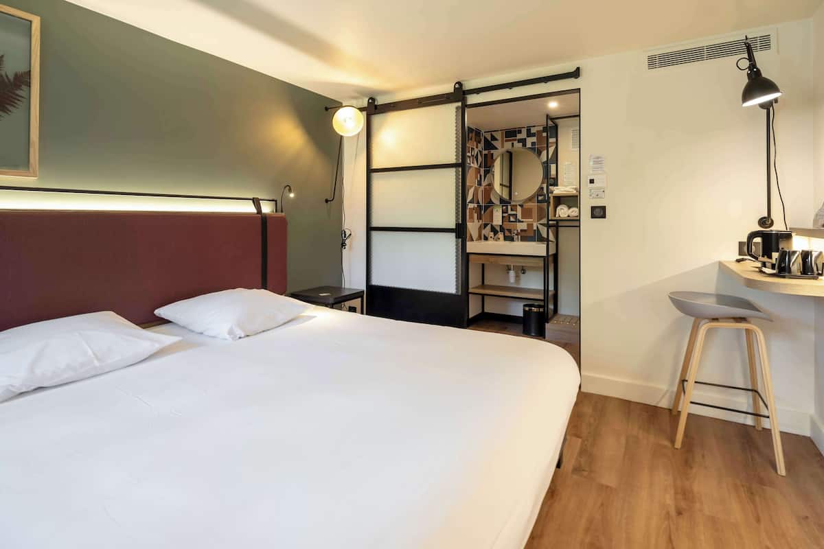 A hotel room with a bed, a desk, a stool, a mirror, and a wall-mounted shelf.