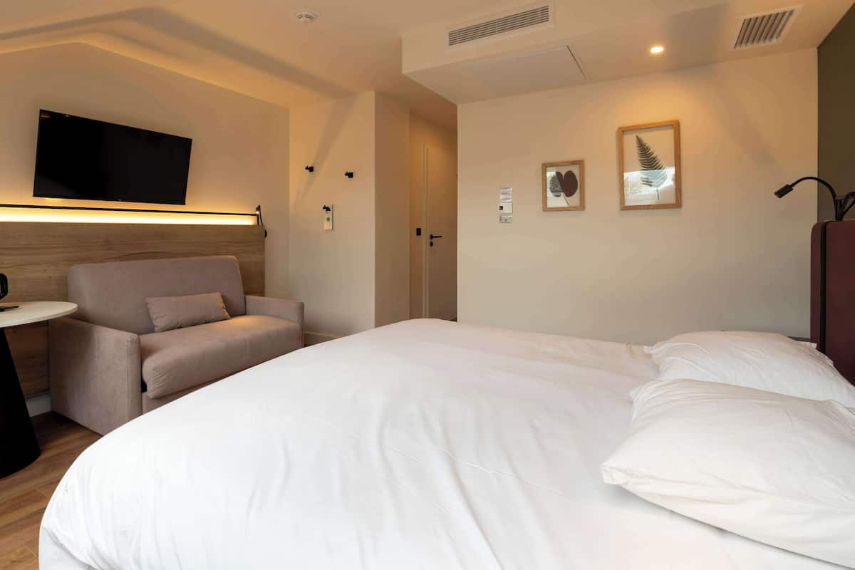 A hotel room with a bed, a chair, a small table, a TV mounted on the wall, and framed pictures on the wall.