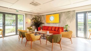 A modern lounge area with a bright orange sofa, yellow chairs, and a large wall-mounted TV. - Campanile NATURE - Metz Sud Jouy aux Arches (Jouy-aux-Arches)