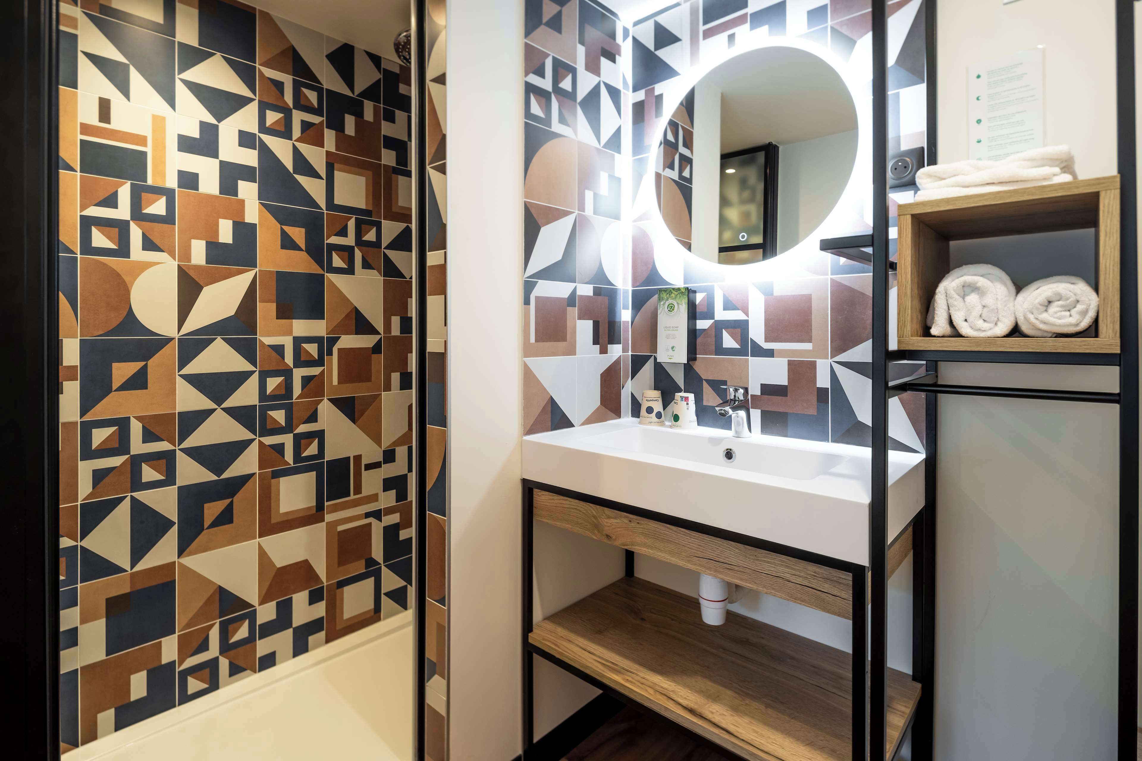 A bathroom with a geometric tile backsplash, a white sink with a mirror, and a wooden shelf with towels.