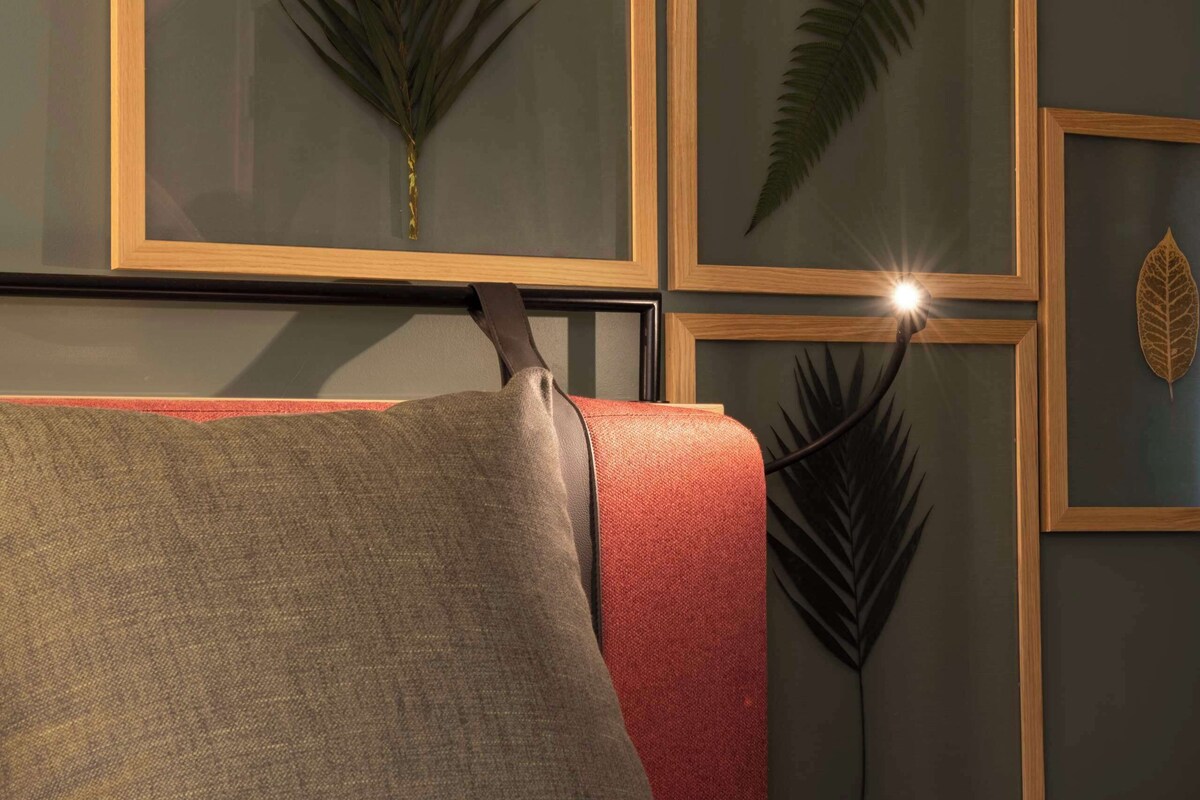 A hotel room with a bed, a red and gray sofa, and framed wall art of palm leaves.