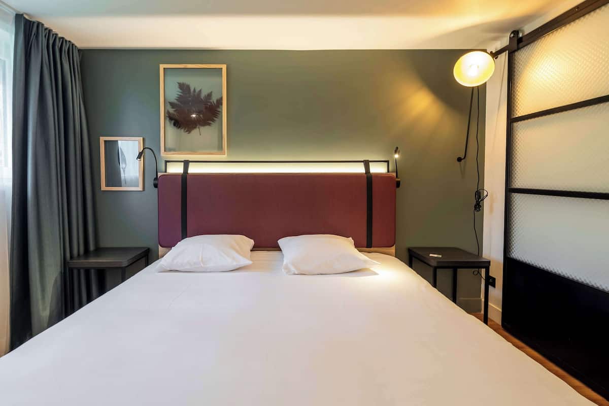 A neatly made bed with white linens and two pillows, a headboard with a red panel, a framed artwork on the wall, a small table beside the bed, and a lamp on the nightstand.