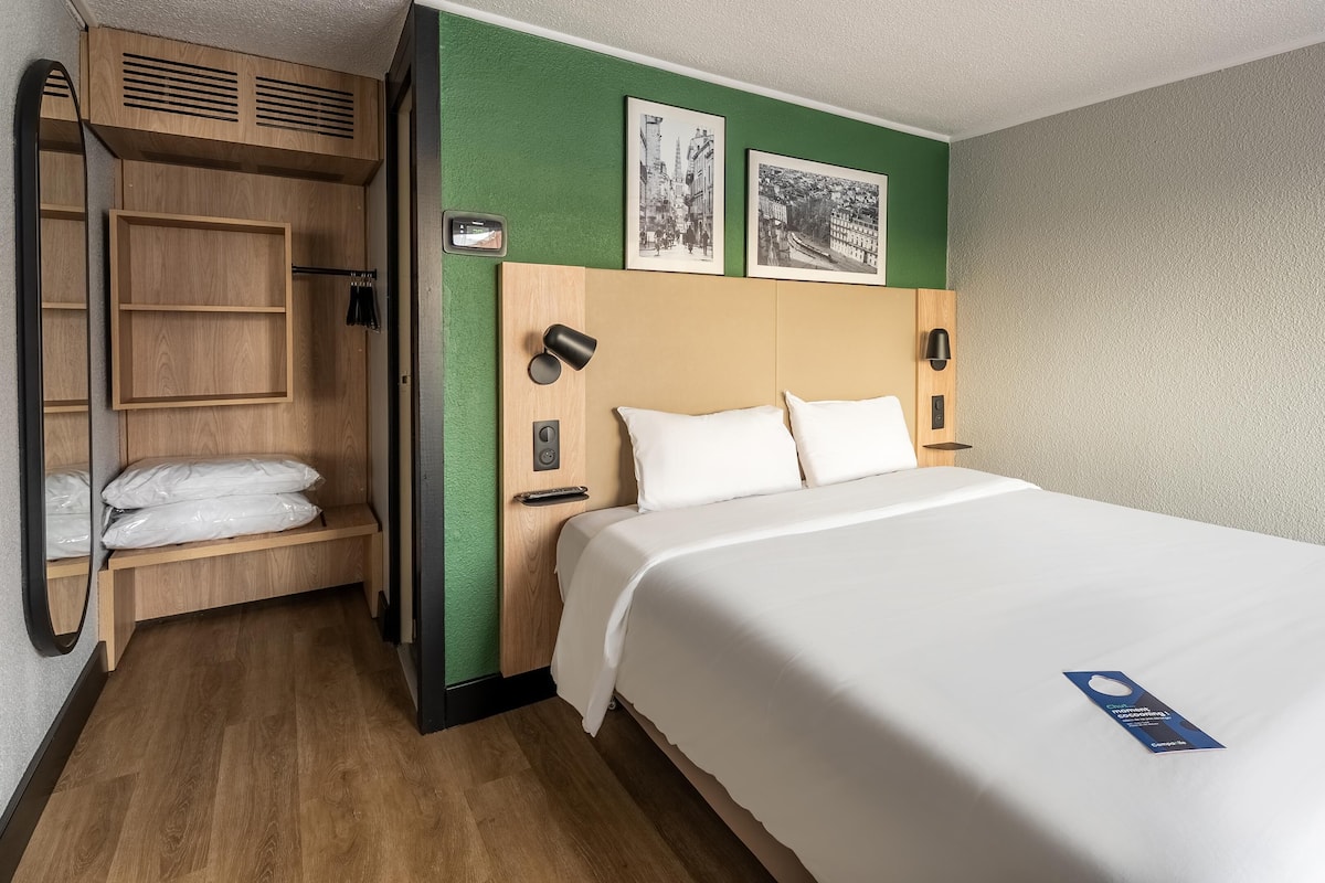 Una habitación de hotel con una cama, un armario de madera, una pared de acento verde y dos cuadros enmarcados.