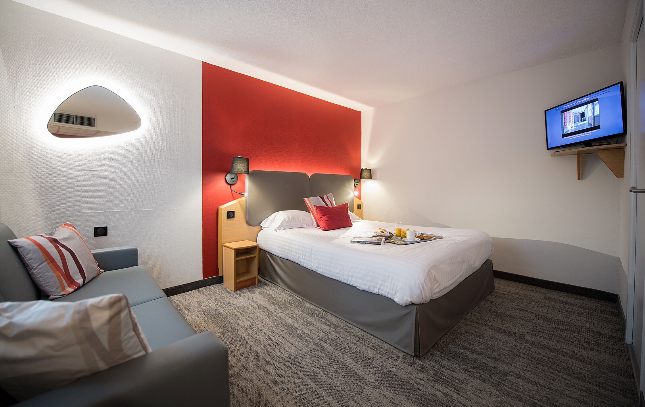 A modern hotel room with a bed, a sofa, a TV, and a red accent wall.