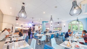 A modern dining area with tables and chairs, a kitchen in the background, and large windows. - Promotel Carros (Carros)