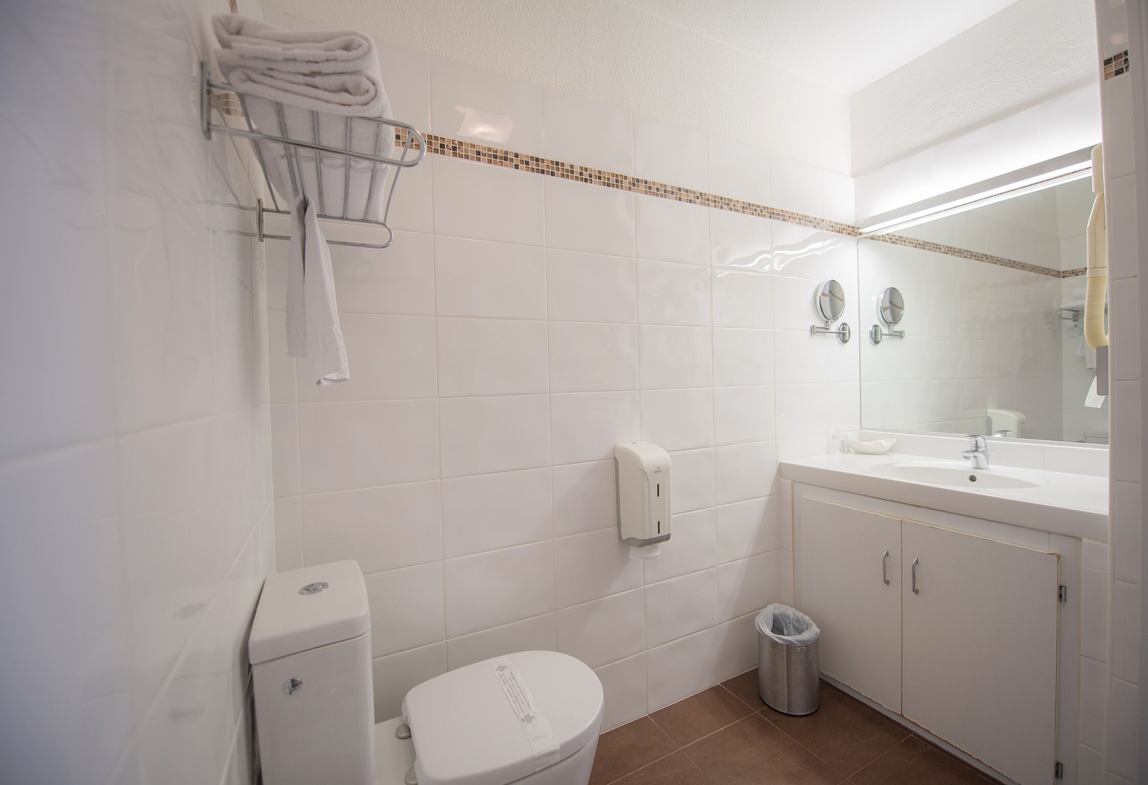 A bathroom with a toilet, a sink, a mirror, a towel rack, and a trash bin.