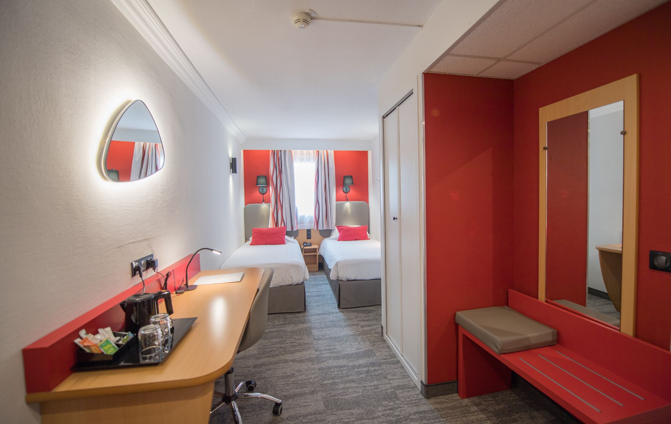 Une chambre d’hôtel équipée d’un bureau, de deux lits, d’une banquette, d’un miroir et d’un mur d’accent de couleur rouge.