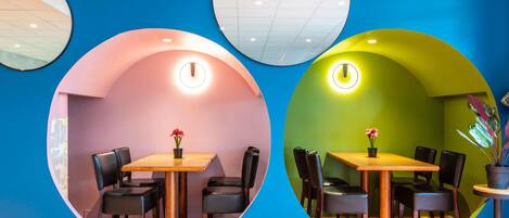 A restaurant with two dining areas separated by a wall with circular cutouts. Each area has a table with chairs and a small plant on the table.