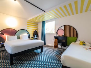 Habitación de hotel con dos camas, un escritorio, una silla, un ventanal con cortinas y suelo estampado.