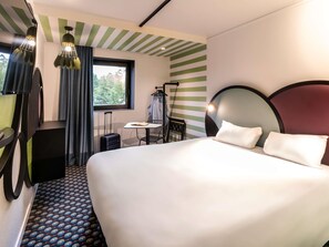 Una habitación de hotel con cama, una mesita, una silla, una maleta y una ventana con cortinas.