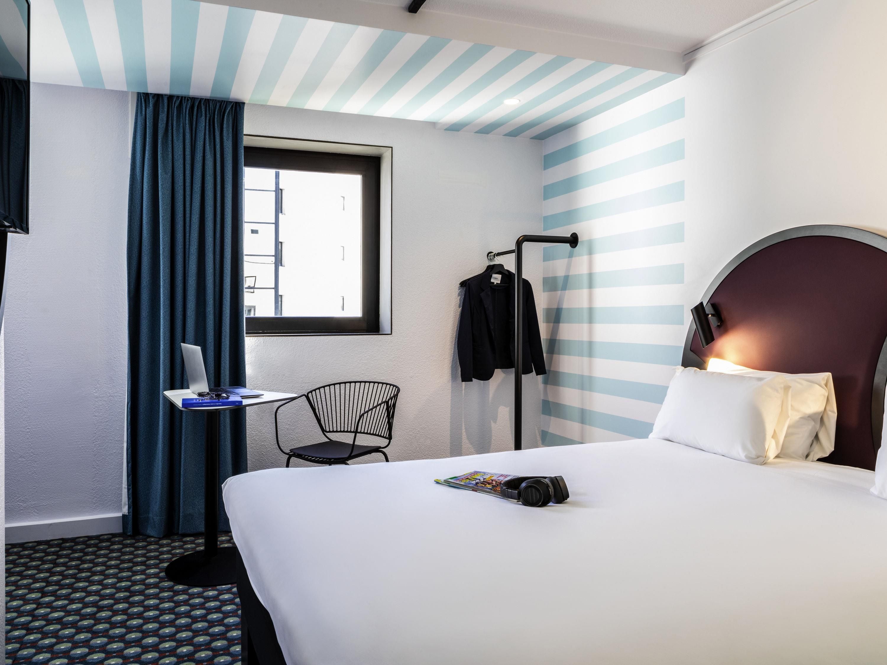 Quarto de hotel com cama, escrivaninha com laptop, cadeira, janela com cortinas e cabideiro.