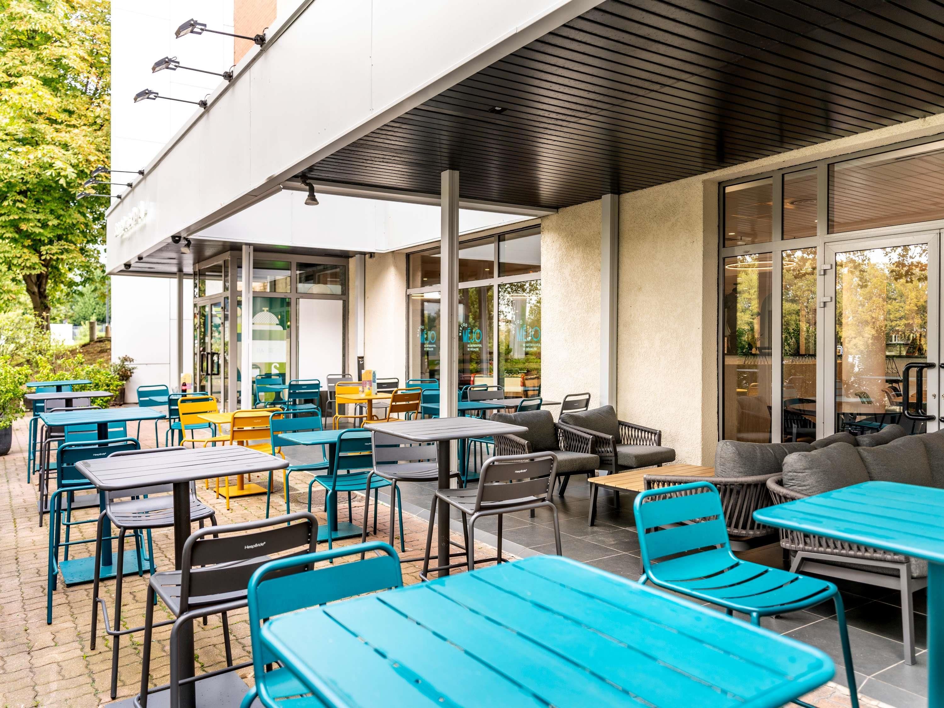 Un café en plein air doté de nombreuses tables et chaises, de grandes fenêtres et d’un espace salon abrité.