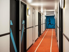 Un couloir avec des portes portant des numéros, un mur bleu orné d’une silhouette en mouvement, et une moquette rouge.