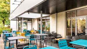 Een terras met meerdere tafels en stoelen, grote ramen en een overdekt zitgedeelte.