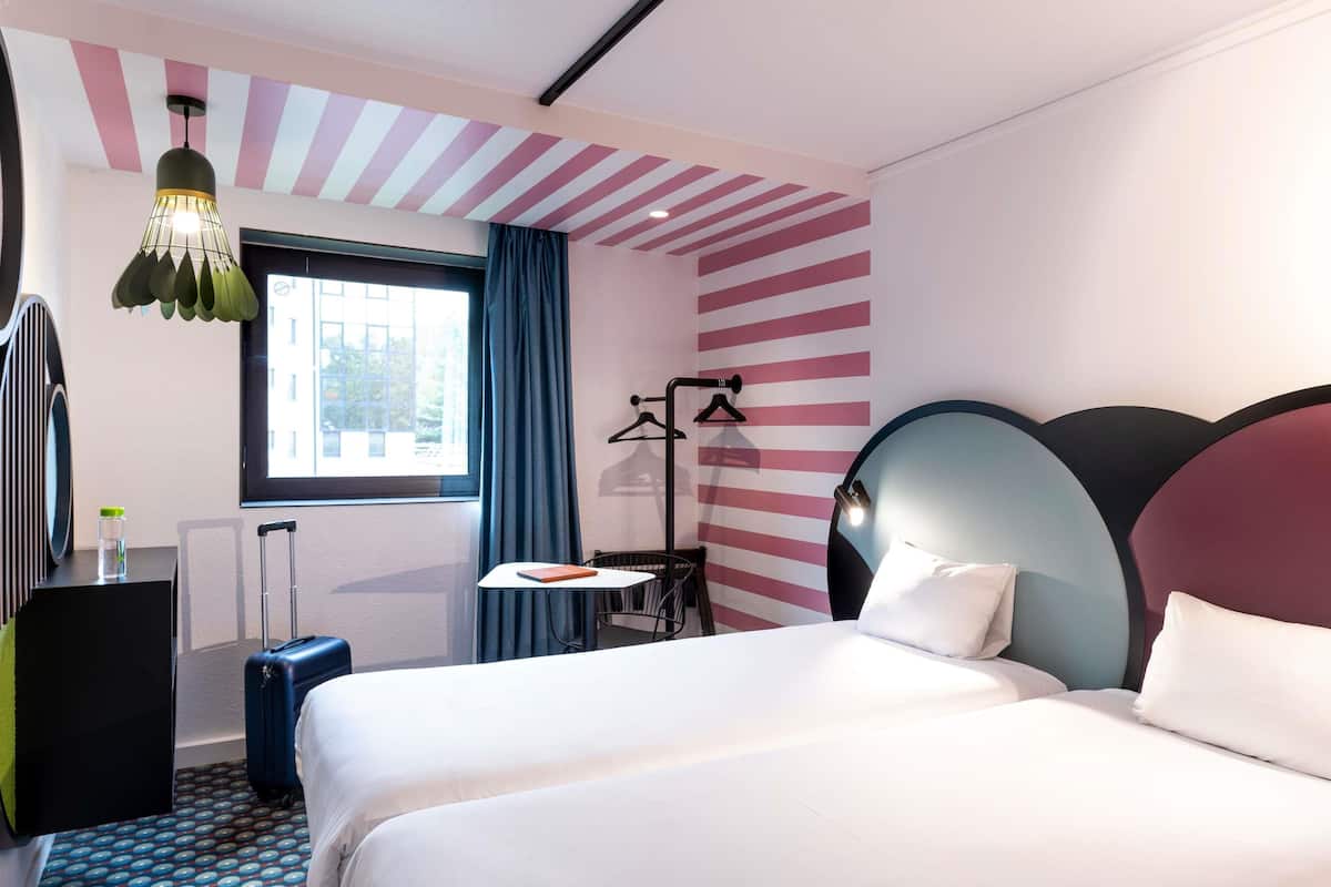 Habitación de hotel con cama, escritorio, silla, lámpara colgante, ventana con cortinas y radiador.