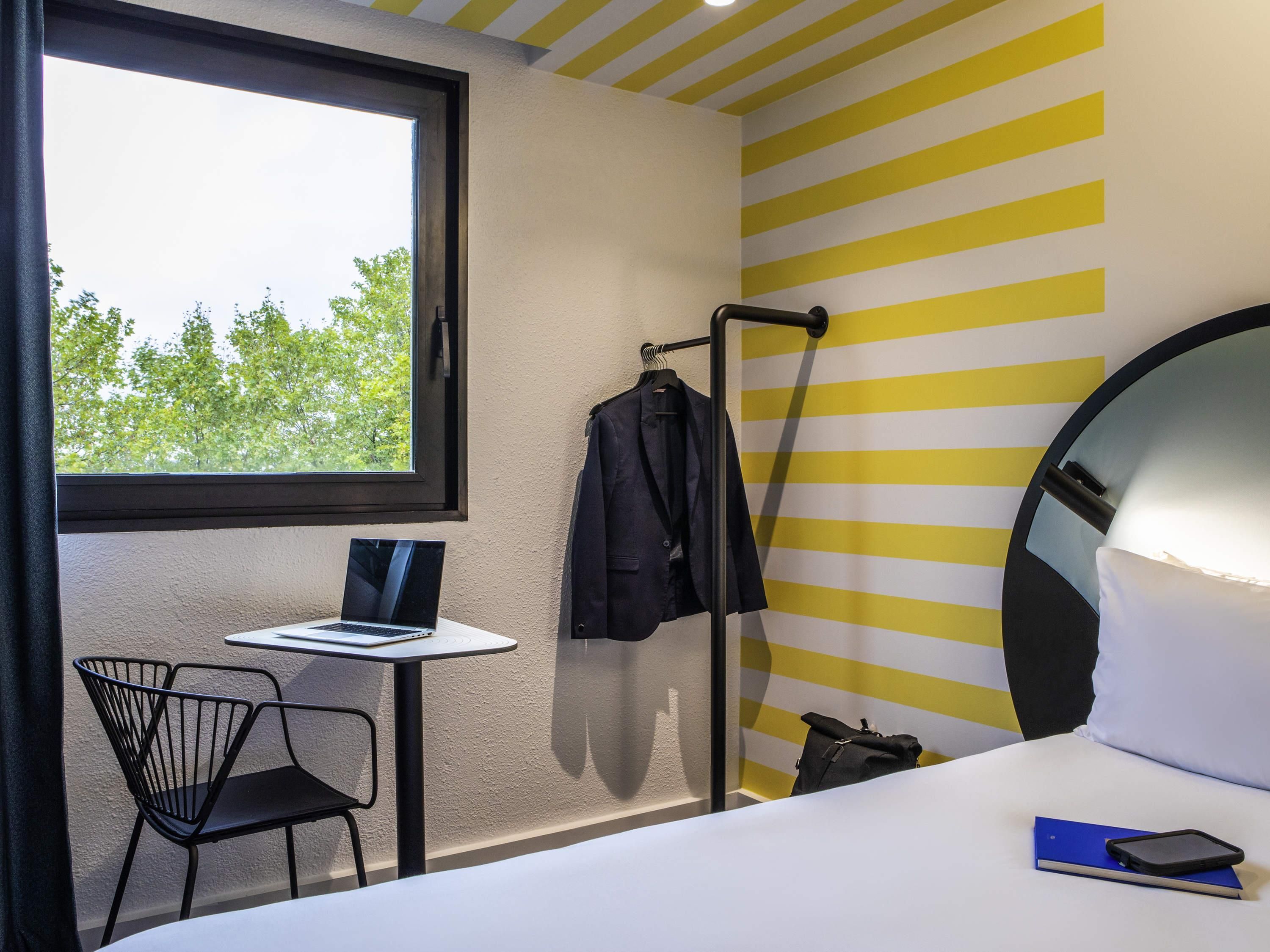 Een hotelkamer met een bed, een bureau met een laptop, een stoel, een kapstok en een raam met uitzicht op bomen.