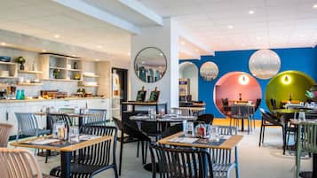 A modern restaurant interior with multiple dining tables and chairs, a kitchen area, and decorative wall elements.
