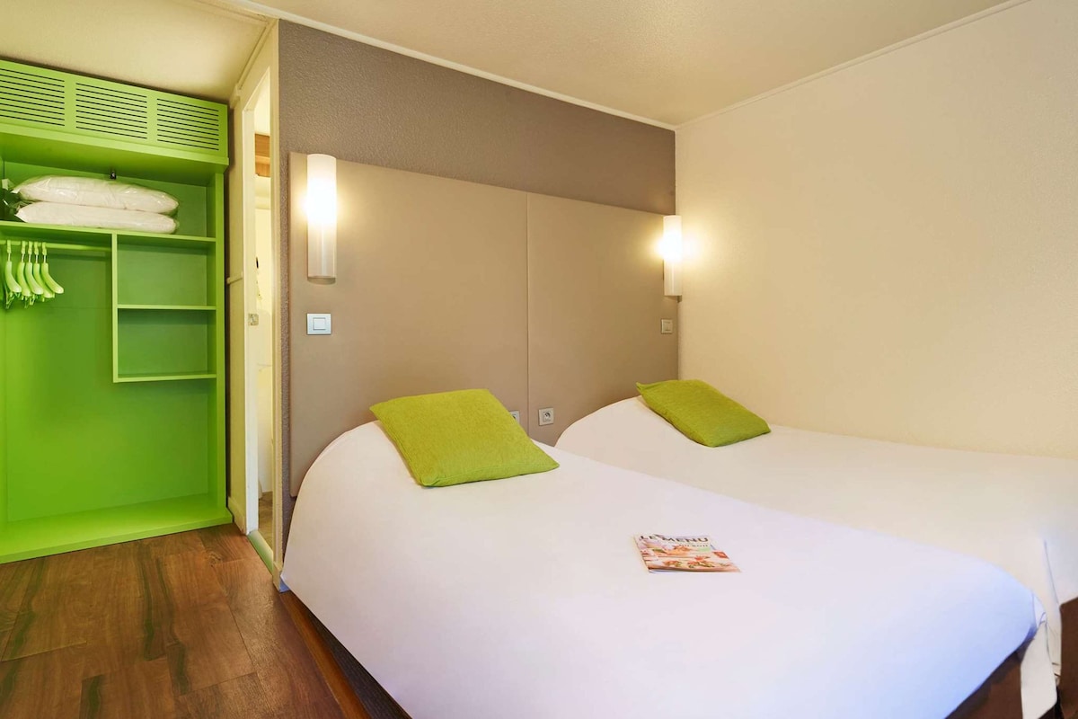 A hotel room with two beds, green pillows, a magazine on the bed, a green wardrobe, and a light switch on the wall.