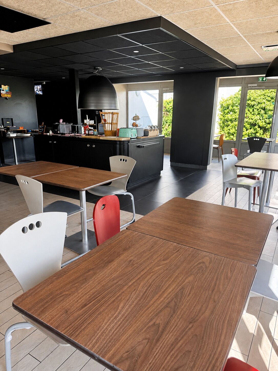 Een modern café met houten tafels en stoelen, een zwarte toonbank met keukenapparatuur, en grote ramen met uitzicht op groen.