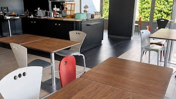 Een modern café met houten tafels en stoelen, een zwarte toonbank met keukenapparatuur, en grote ramen met uitzicht op groen.