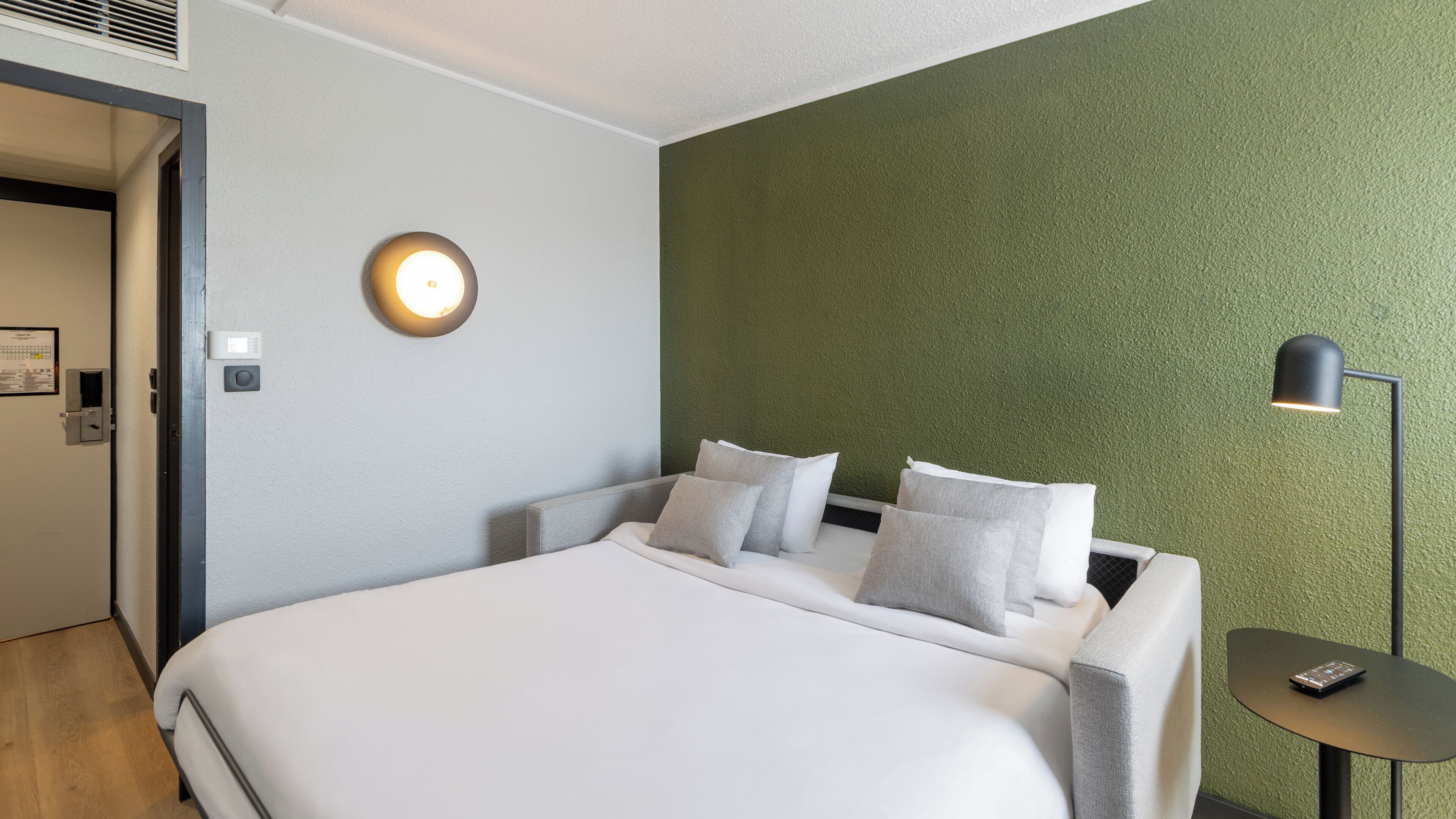 Een hotelkamer met een bed, een nachtkastje, een lamp en een wandlamp.