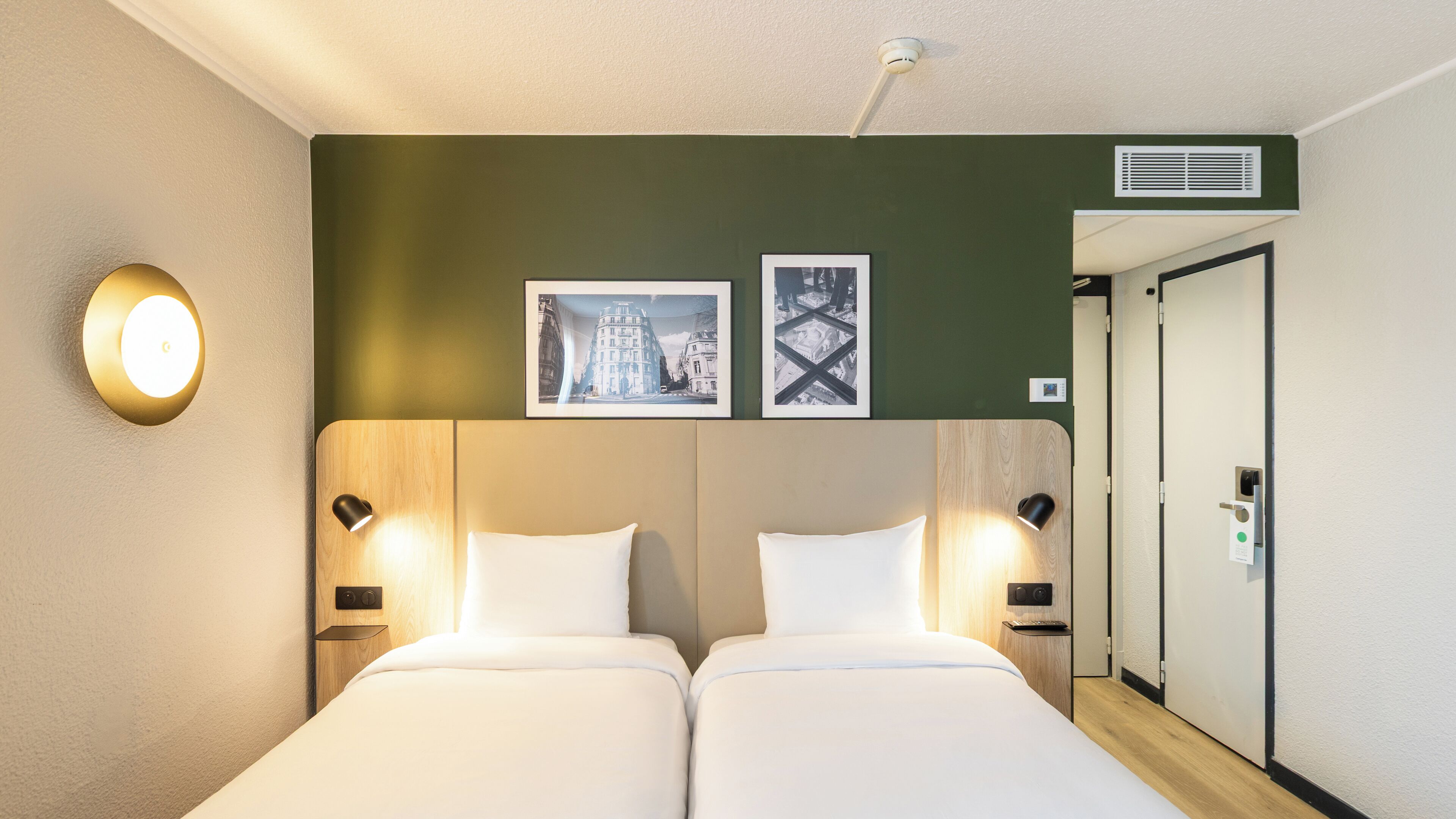 Habitación de hotel con dos camas, una lámpara fijada a la pared, dos cuadros enmarcados, una puerta y una rejilla de ventilación en la pared.