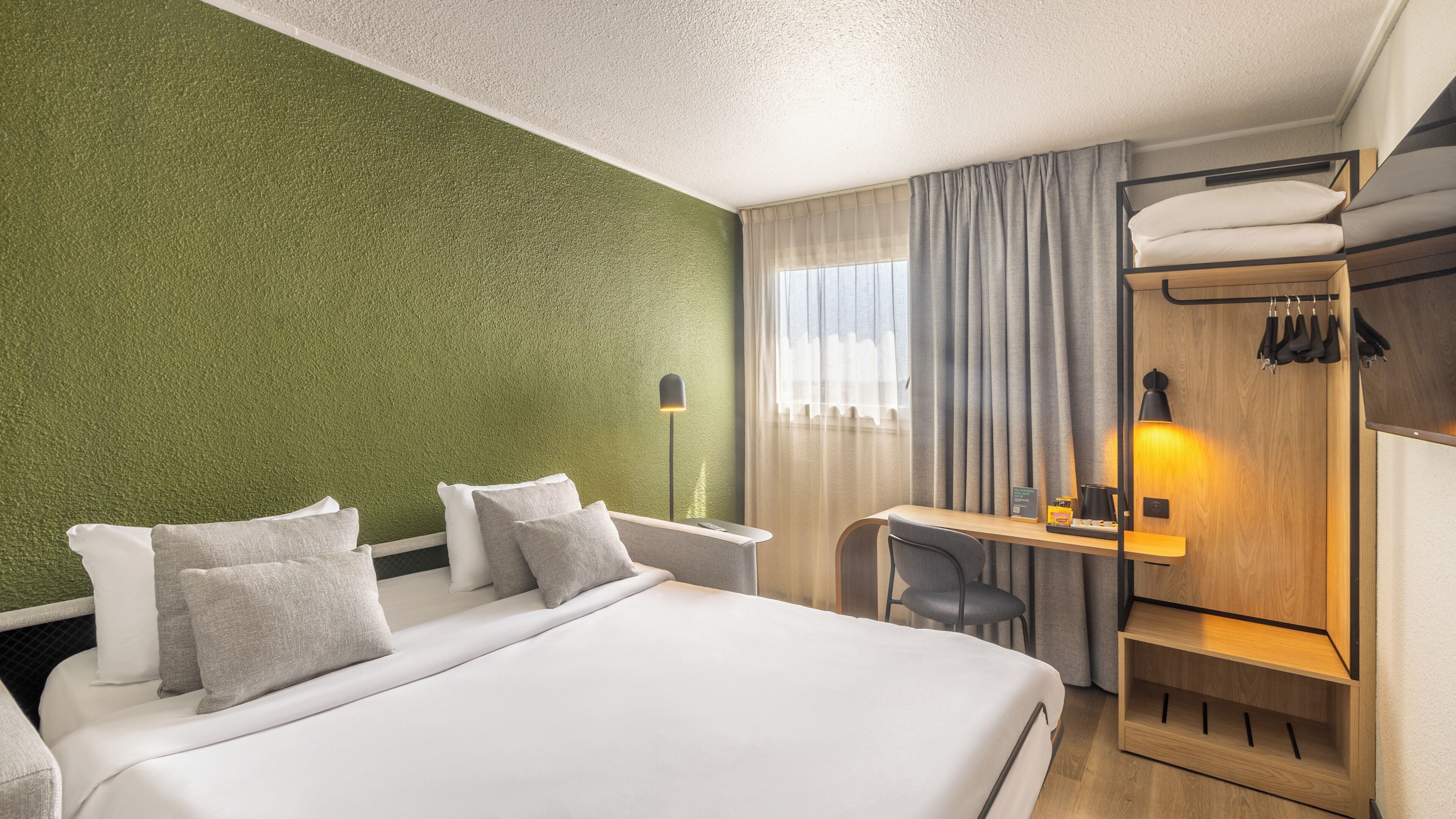 A hotel room with a bed, a desk, a chair, a wardrobe, and a lamp.