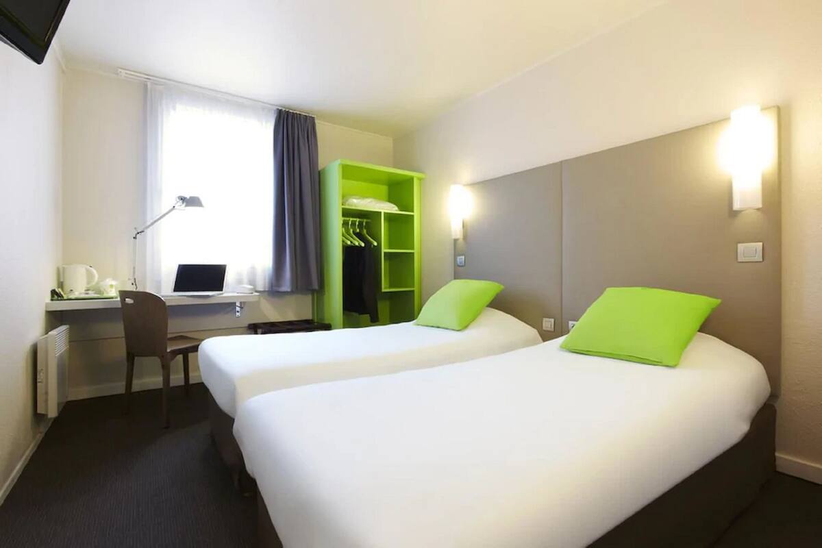 A hotel room with two beds, a desk with a computer, a chair, and a green wardrobe.
