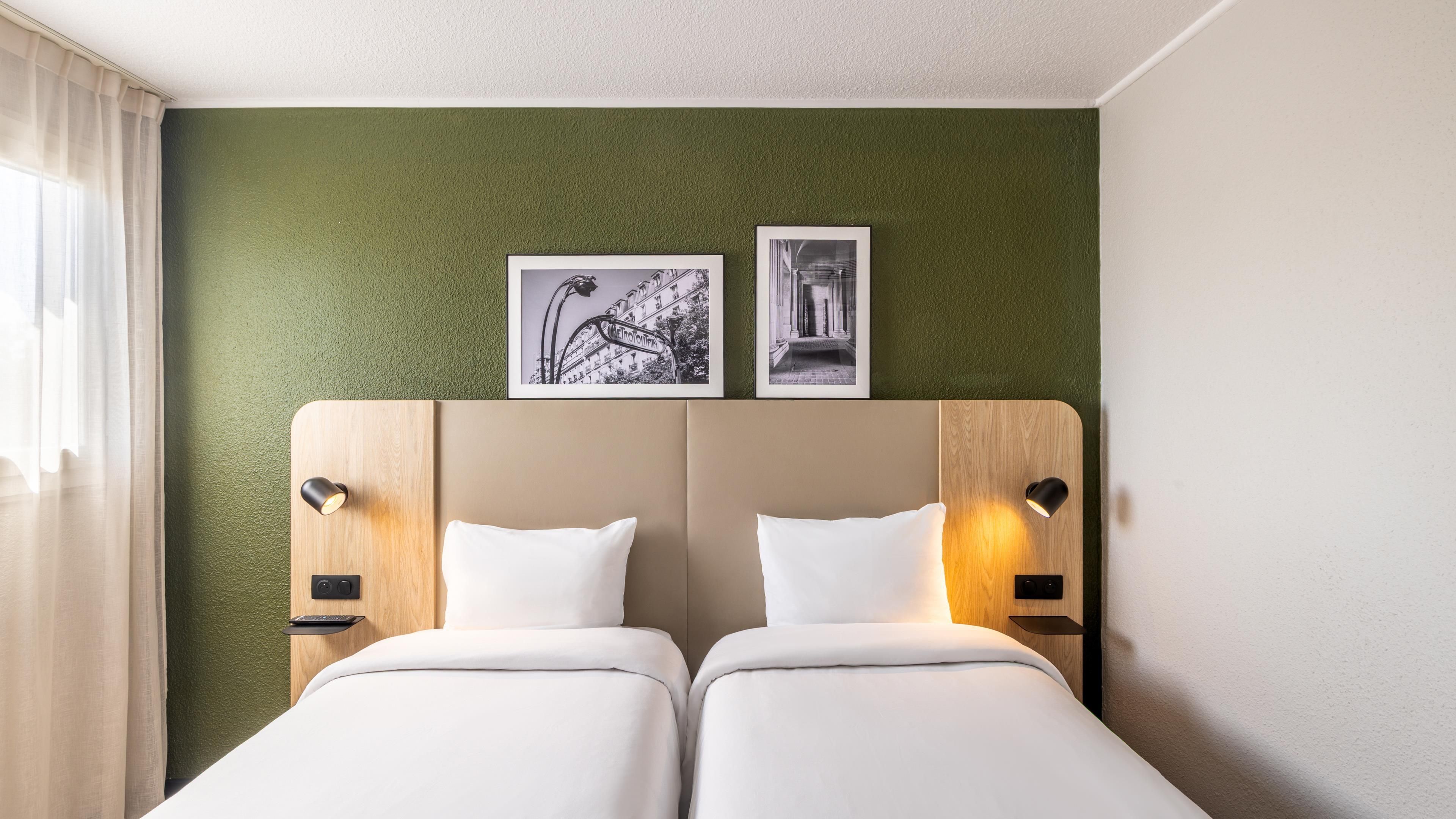 Une chambre d’hôtel avec deux lits, un mur vert à texture et deux tableaux encadrés au-dessus des lits.