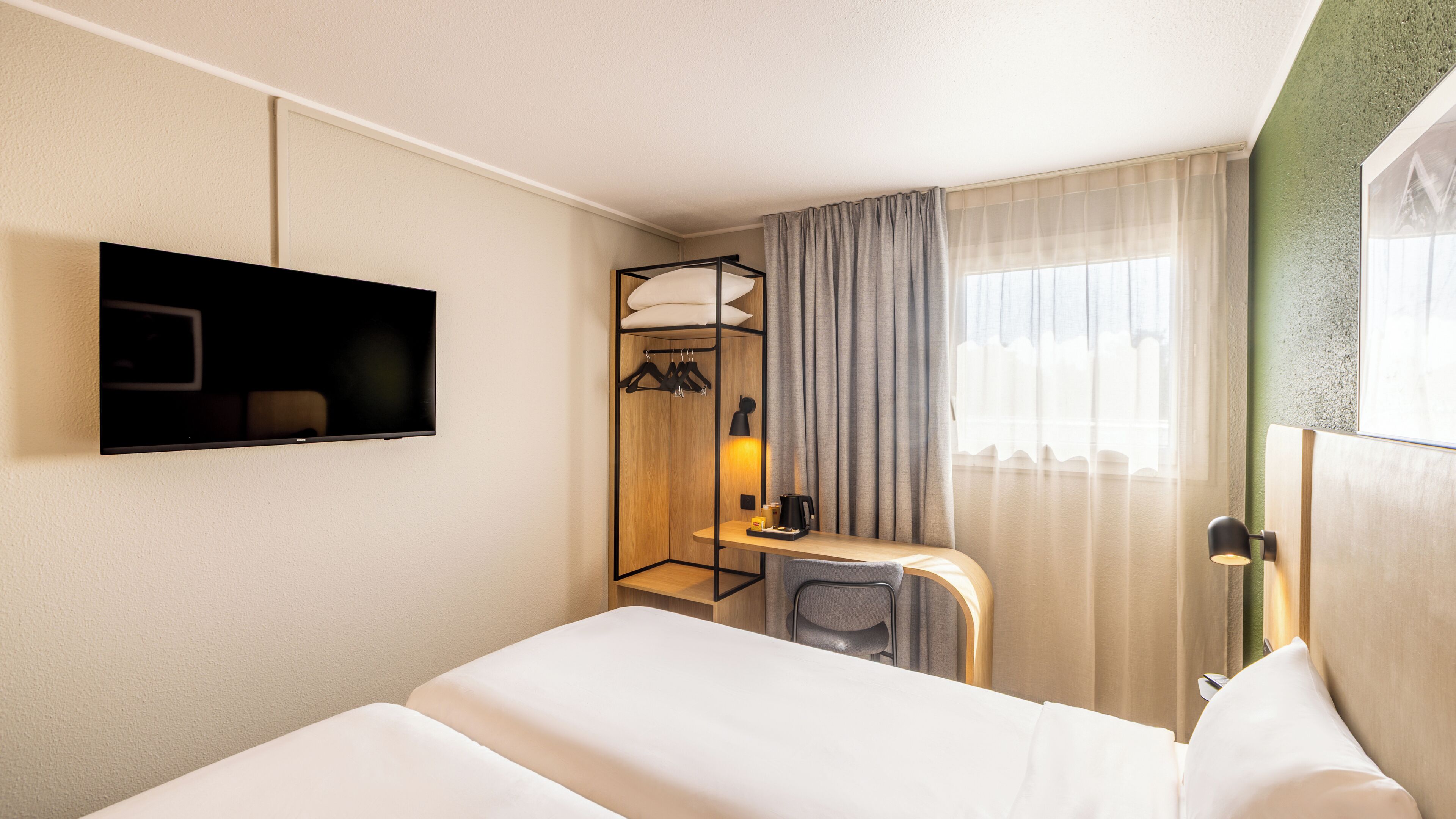 A hotel room with a bed, a TV mounted on the wall, a desk with a lamp, and a window with curtains.