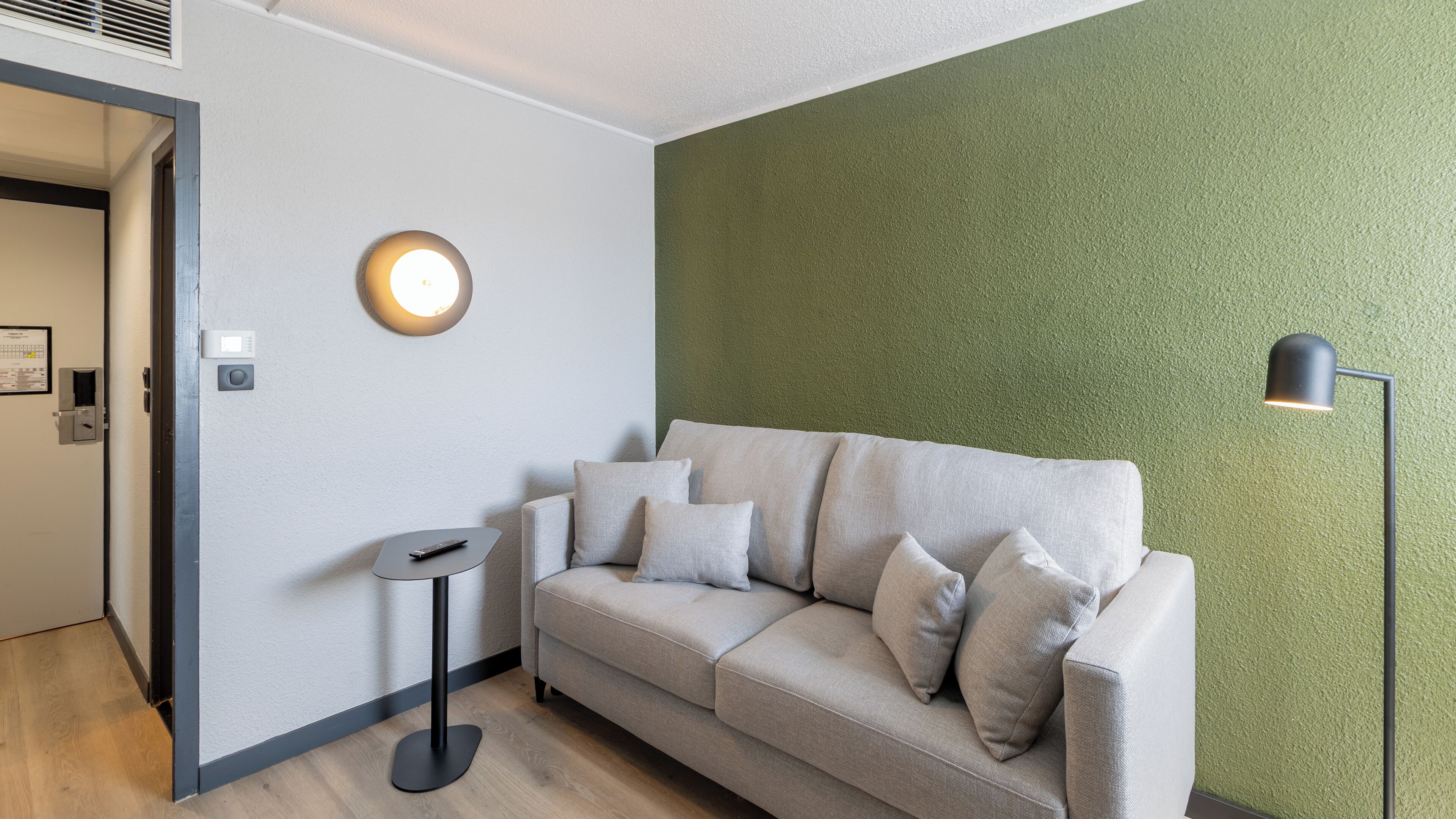 Un moderno salón con un sofá beige, una pequeña mesita auxiliar, una lámpara de pared y una pared verde con textura.