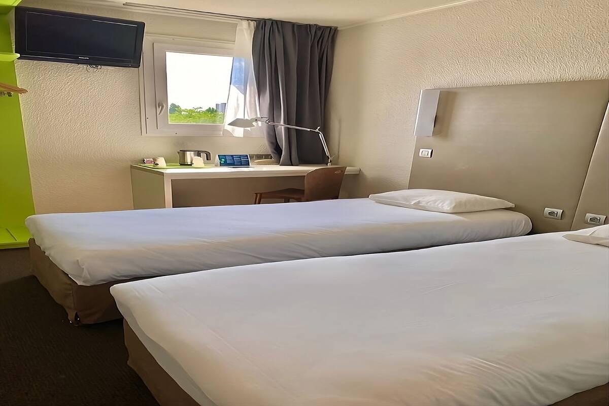 A hotel room with two beds, a desk with a computer, a TV mounted above the desk, and a window with curtains.