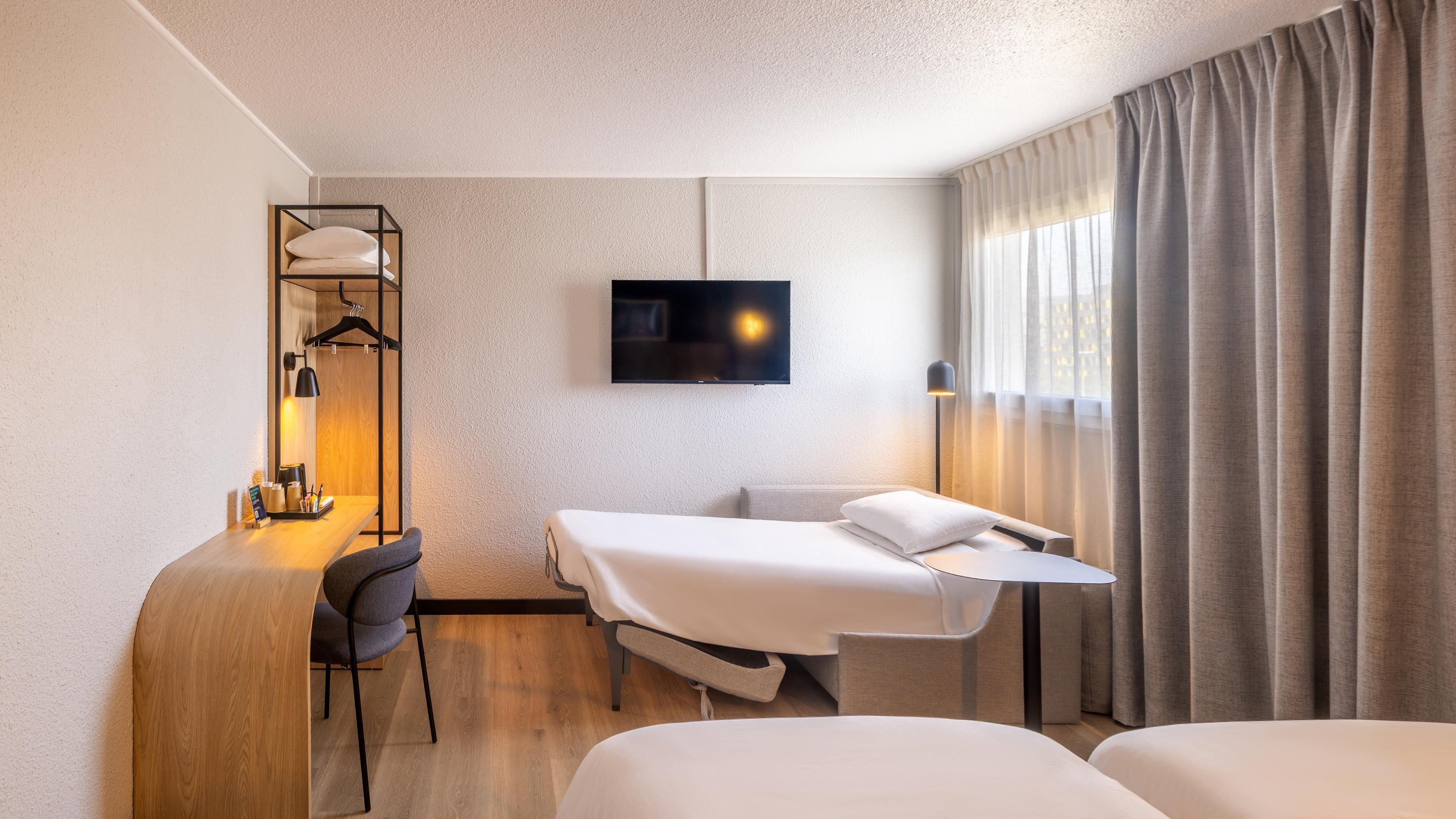 A hotel room with a bed, a desk with a chair, a TV, and a window with curtains.