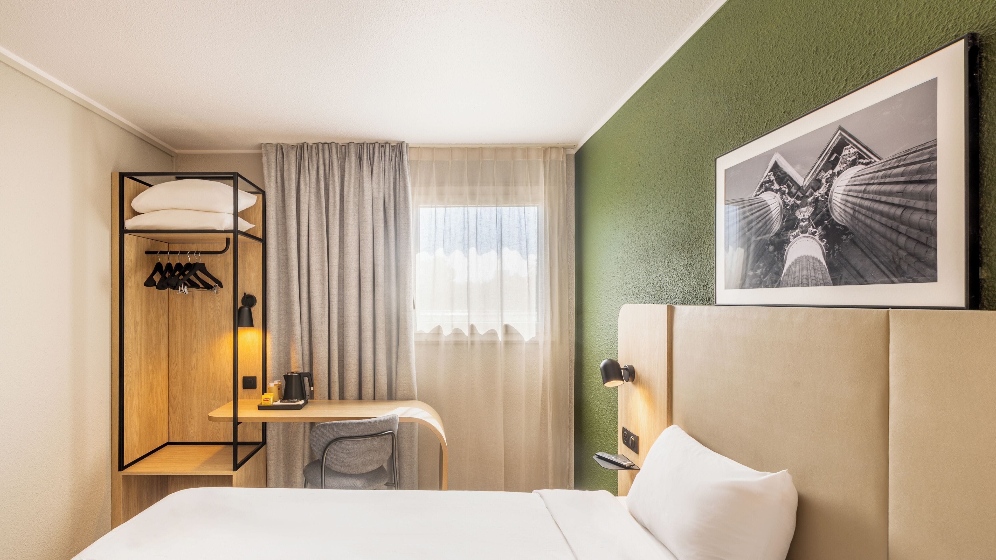 A hotel room with a bed, a desk, a chair, a wardrobe, a window with curtains, a wall-mounted picture, and a wall-mounted lamp.