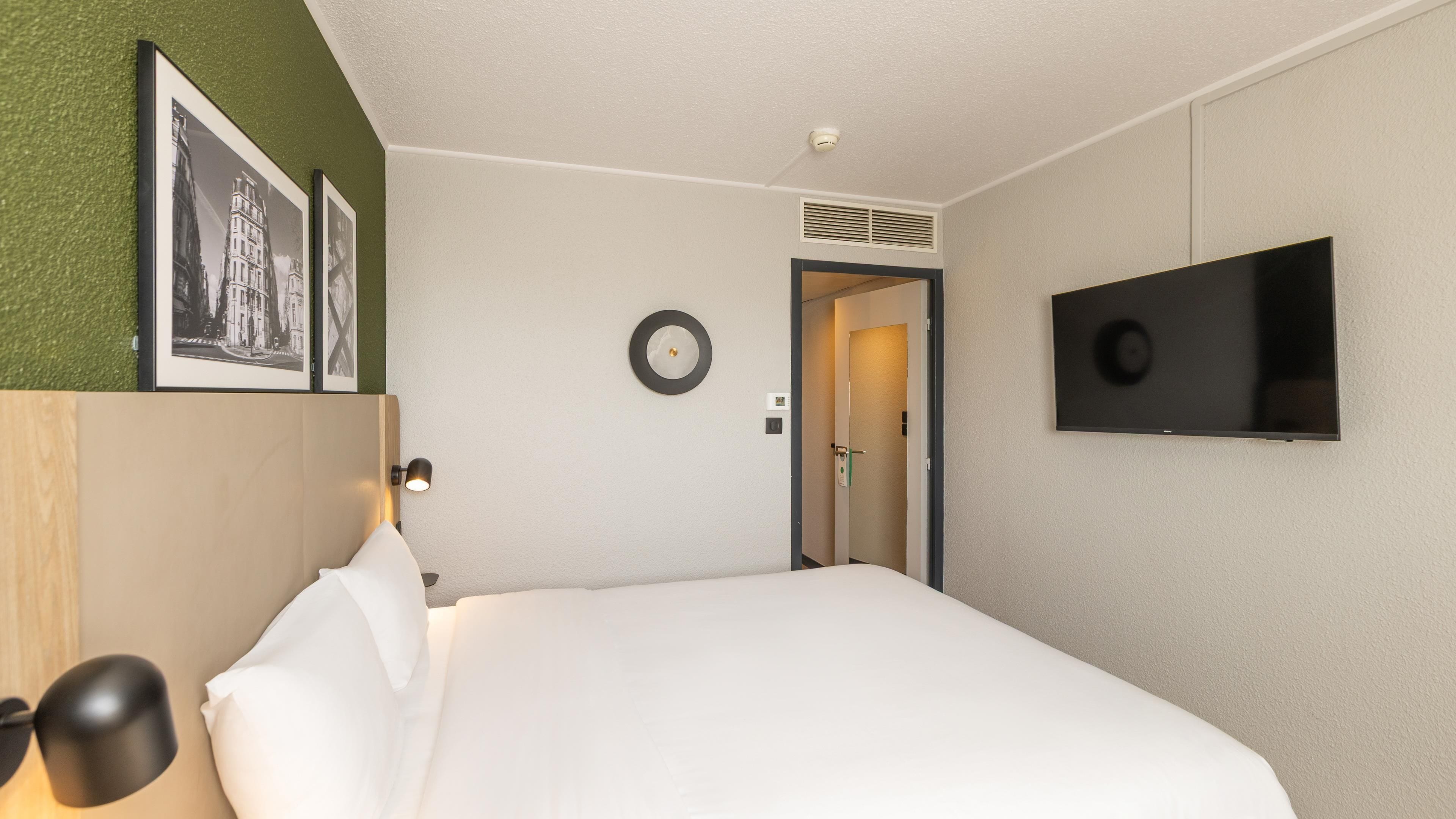 A hotel room with a bed, a flat-screen TV, a wall-mounted clock, and framed pictures.