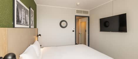 A hotel room with a bed, a flat-screen TV, a wall-mounted clock, and framed pictures.