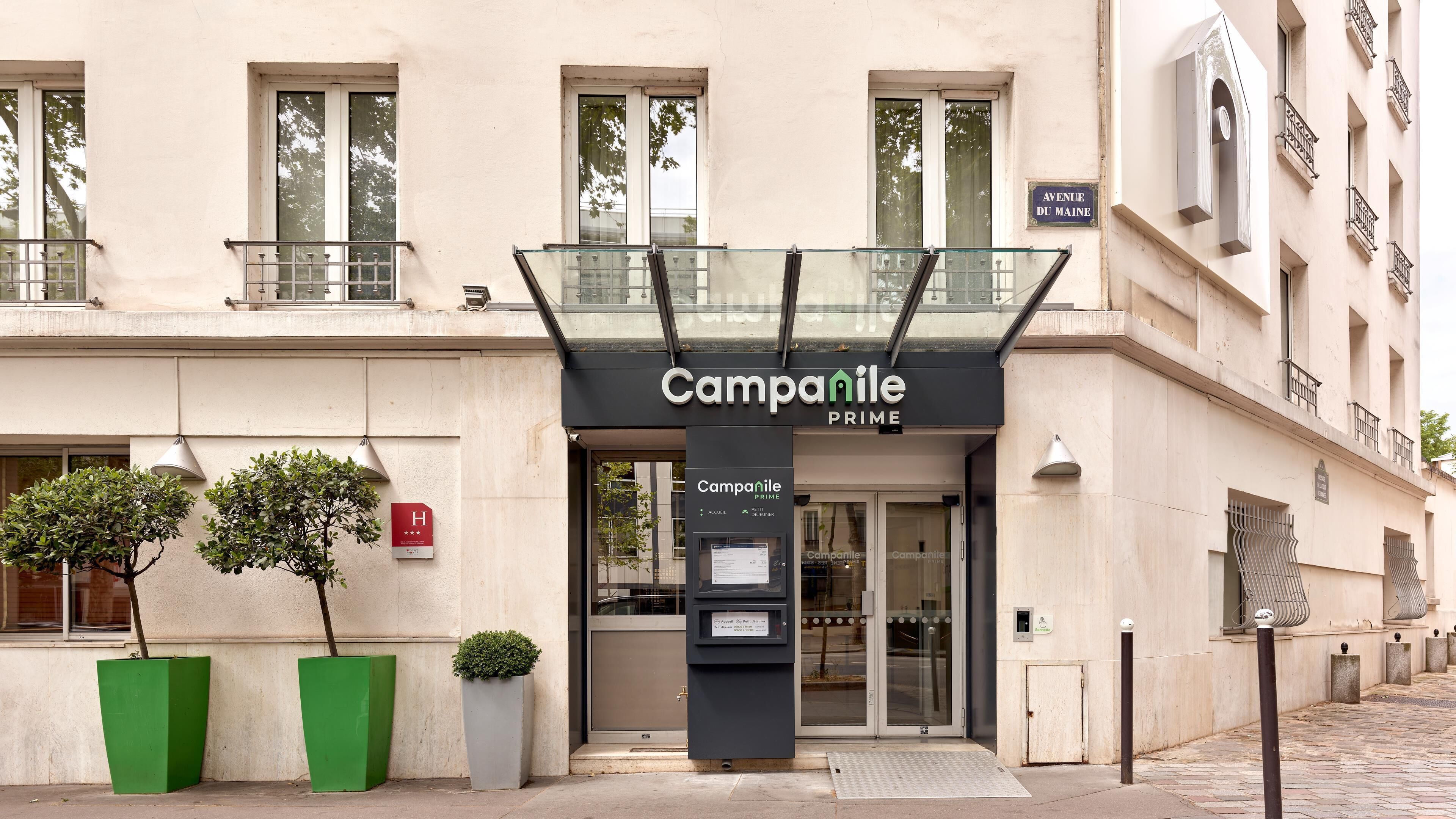 A hotel entrance with a glass canopy, a sign reading 'Campanile PRIME', and a small outdoor seating area with potted plants.