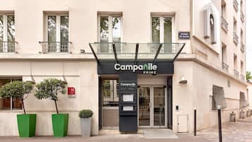 A hotel entrance with a glass canopy, a sign reading 'Campanile PRIME', and a small outdoor seating area with potted plants.