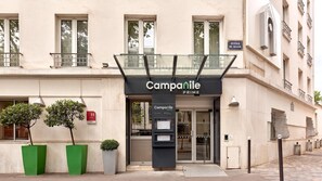 Entrada de un hotel con un toldo de cristal, un cartel que dice 'Campanile PRIME' y una pequeña zona de estar al aire libre con plantas en macetas.