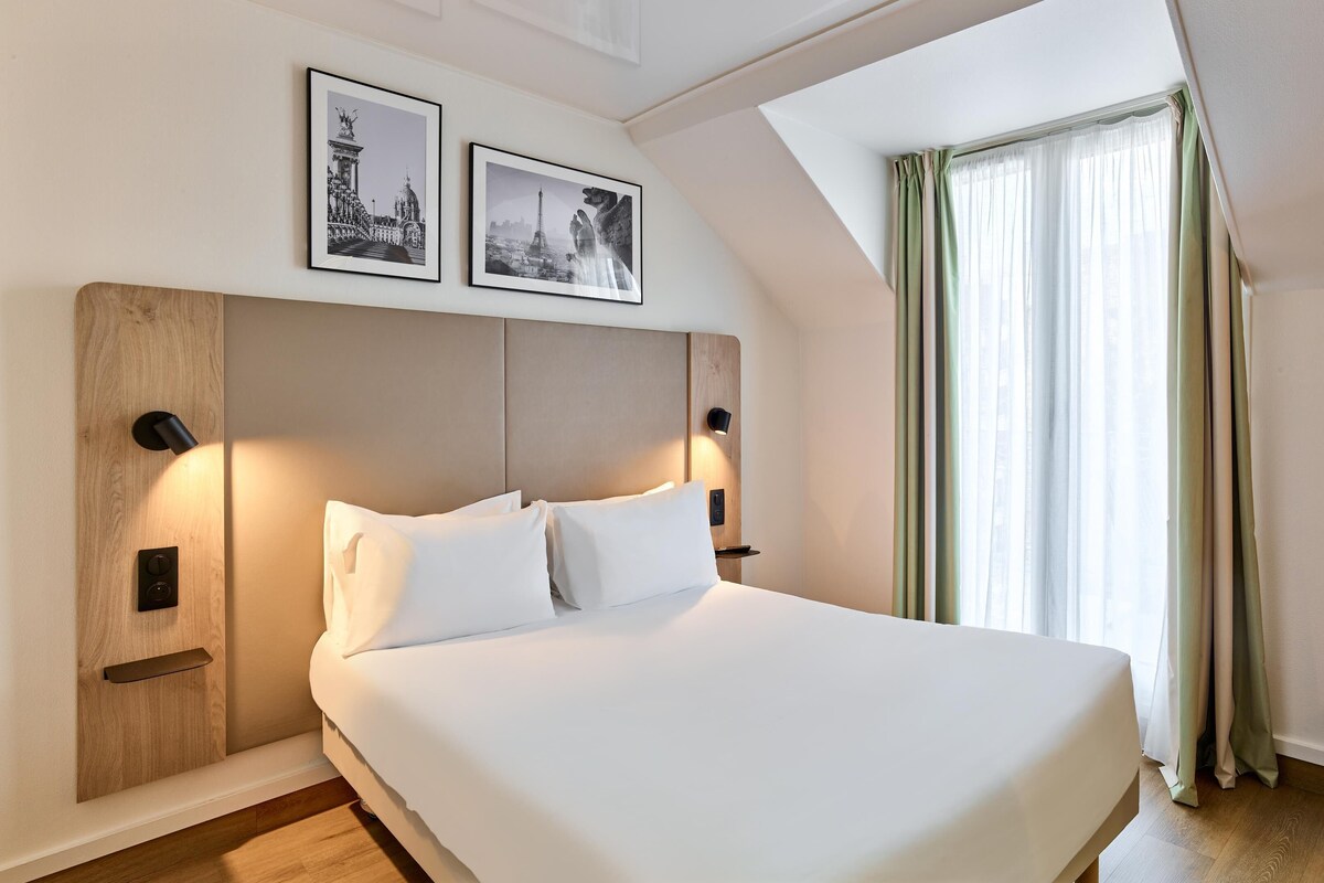 A hotel room with a bed, a nightstand, a wall-mounted lamp, a window with curtains, and two framed pictures on the wall.